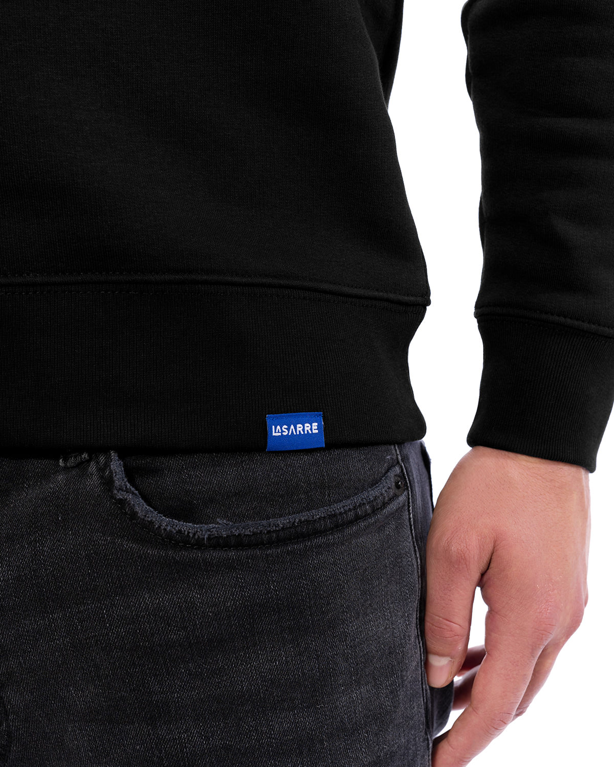 Eleganter Sweater in Schwarz, Detailansicht. Zu sehen ist die linke Hüft- und Bauchpartie des Models und damit das vordere blaue Saum-Schildchen mit LASARRE Schriftzug in Großaufnahme.