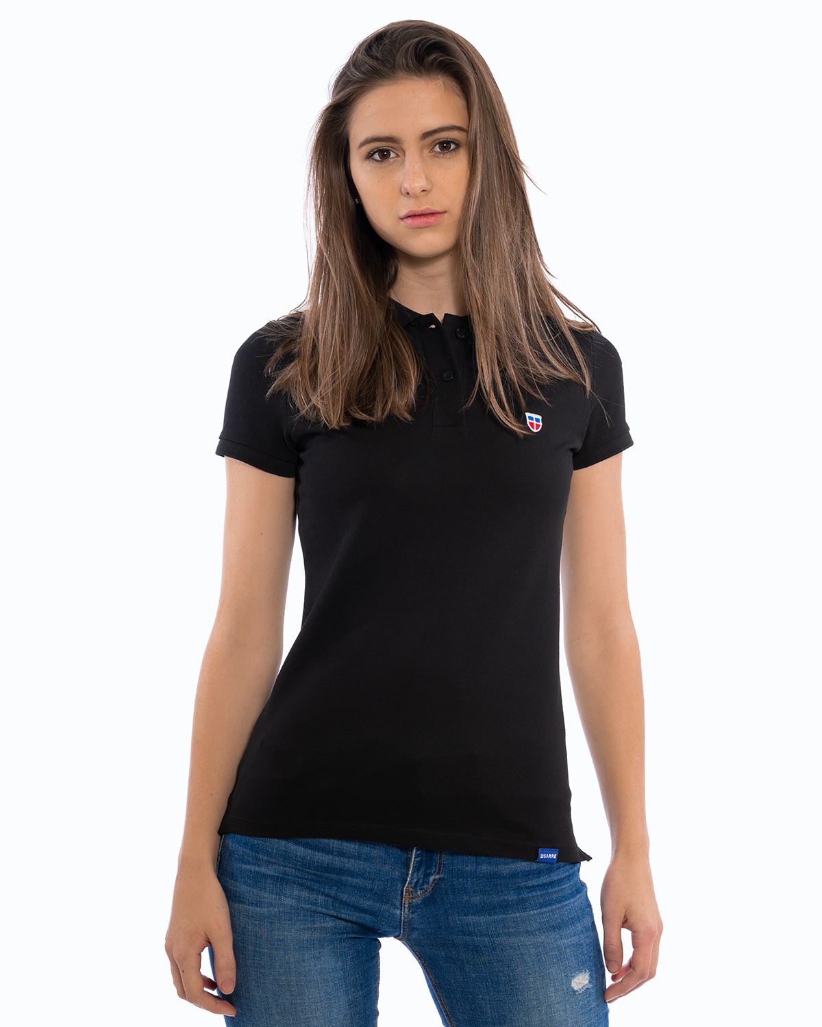 Junges weibliches Model in entspannter Pose im schwarzen Ladies-Polo-Shirt der Saarland-Marke LA SARRE. Auffällig sind das blaue LA SARRE Saumlabel und das Sarre-Wappen als Stickerei auf der Brust.