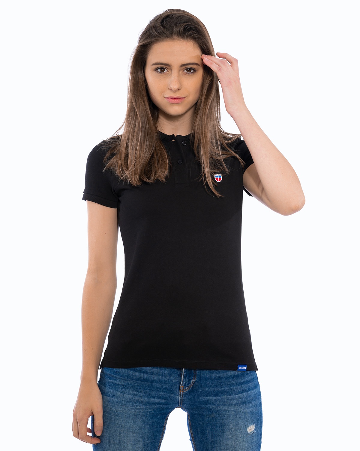 Weibliches Model in lockerer Pose im LA SARRE Ladies-Polo-Shirt. Frontal-Ansicht des Oberteils der Saarland Marke in der Farbe Schwarz mit blauem LA SARRE Saumlabel und dem Sarre-Wappen als Stickerei auf der linken Brust.
