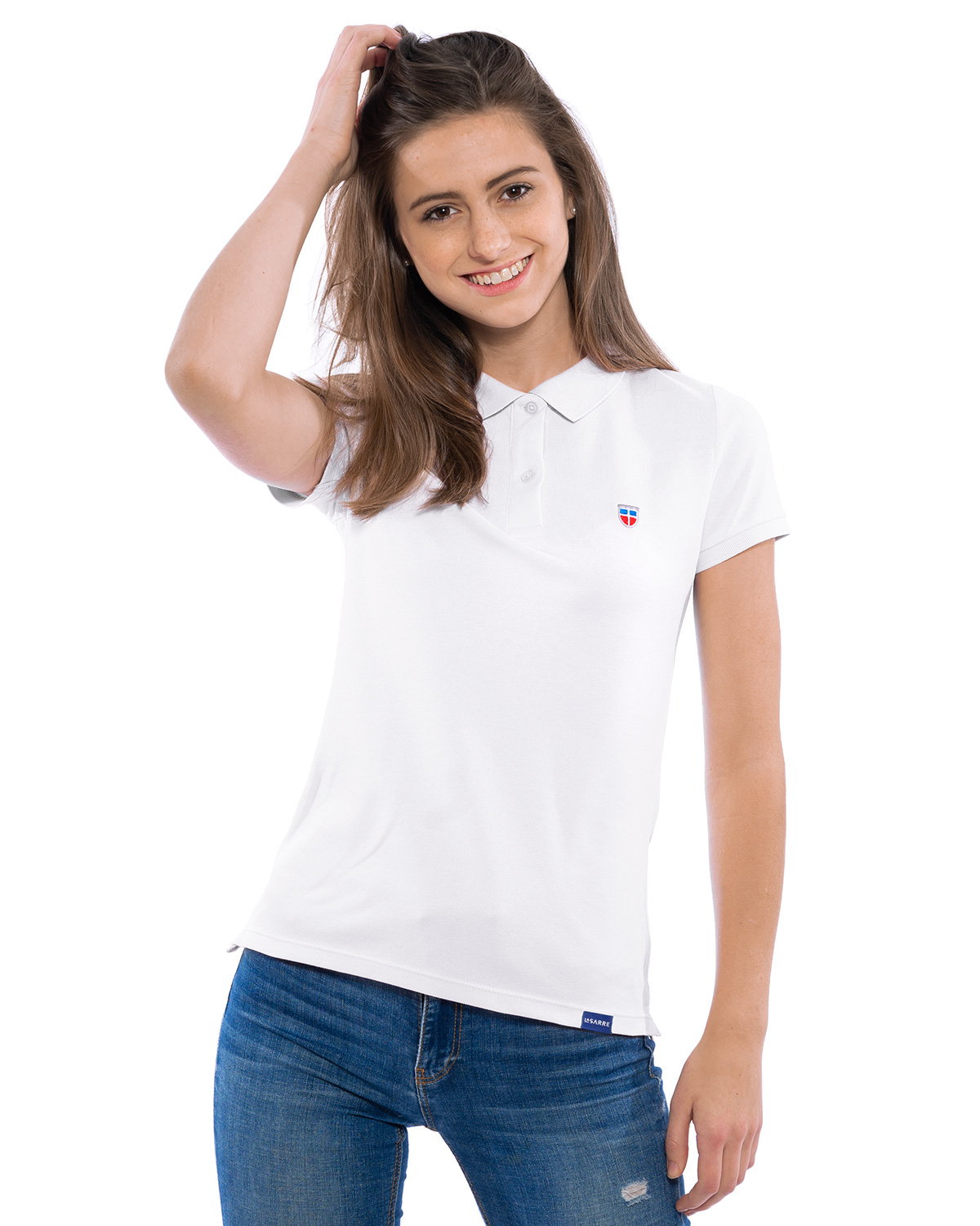 Junge Frau in lockerer Pose im LA SARRE Ladies-Polo-Shirt. Frontal-Ansicht des Oberteils der Saarland Marke in der Farbe Weiß mit blauem LA SARRE Saumlabel und dem Sarre-Wappen als Stickerei auf der Brust.