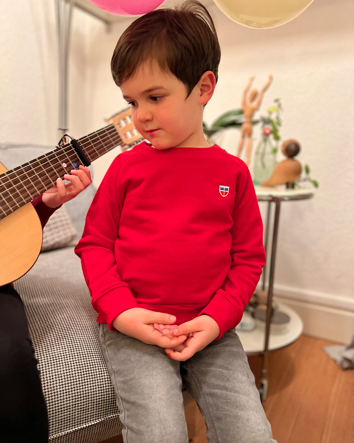 Wir sehen Anton, 5 Jahre alt in unserem Kinder Pullover Tom mit Saarland Wappen auf der linken Brust. Er schaut beeindruckt seiner Schwester beim Gitarrenspielen zu.