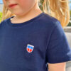 Wir sehen das Sarre-Wappen auf Selmas navy-blauem T-Shirt in Großaufnahme.